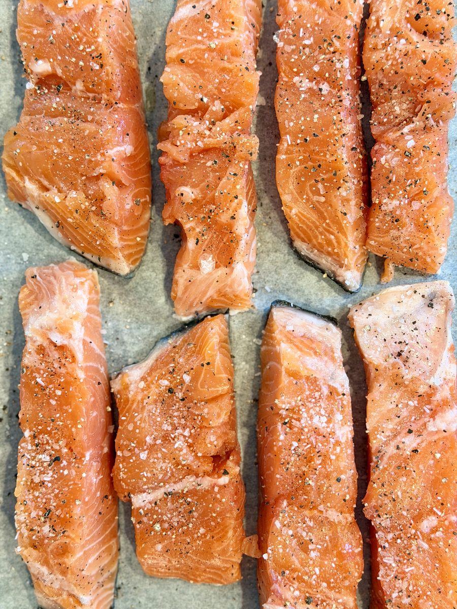 Overhead of eight raw salmon fillets, skin side down and sprinkled with salt, laid in rows on a parchment-lined sheet.