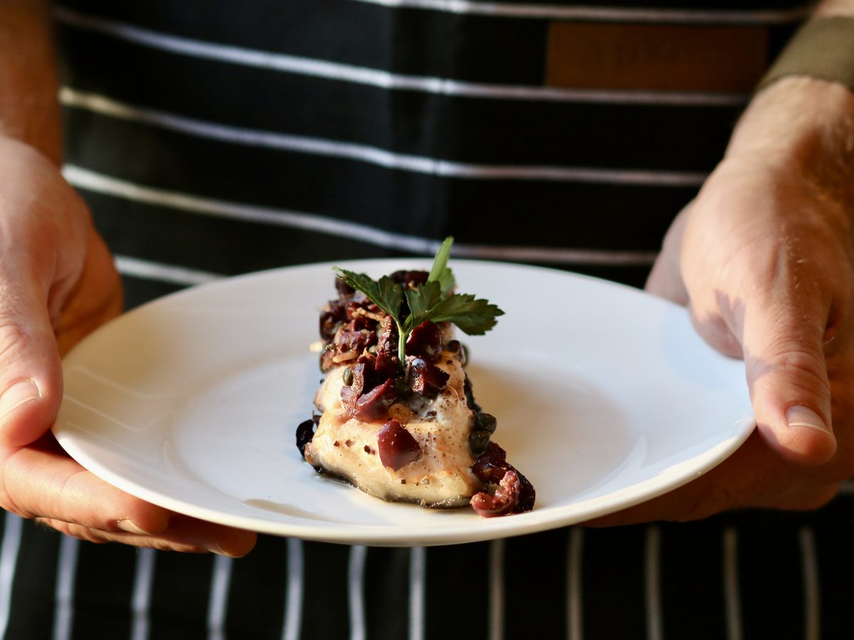 Two hands hold out a white plate bearing a single pale flaky fish fillet topped with chopped olives, capers, red onion and tomato