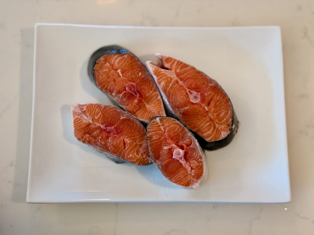 Three raw salmon steaks with skin edges and center bone visible, arranged on a white rectangular platter on a marble counter.