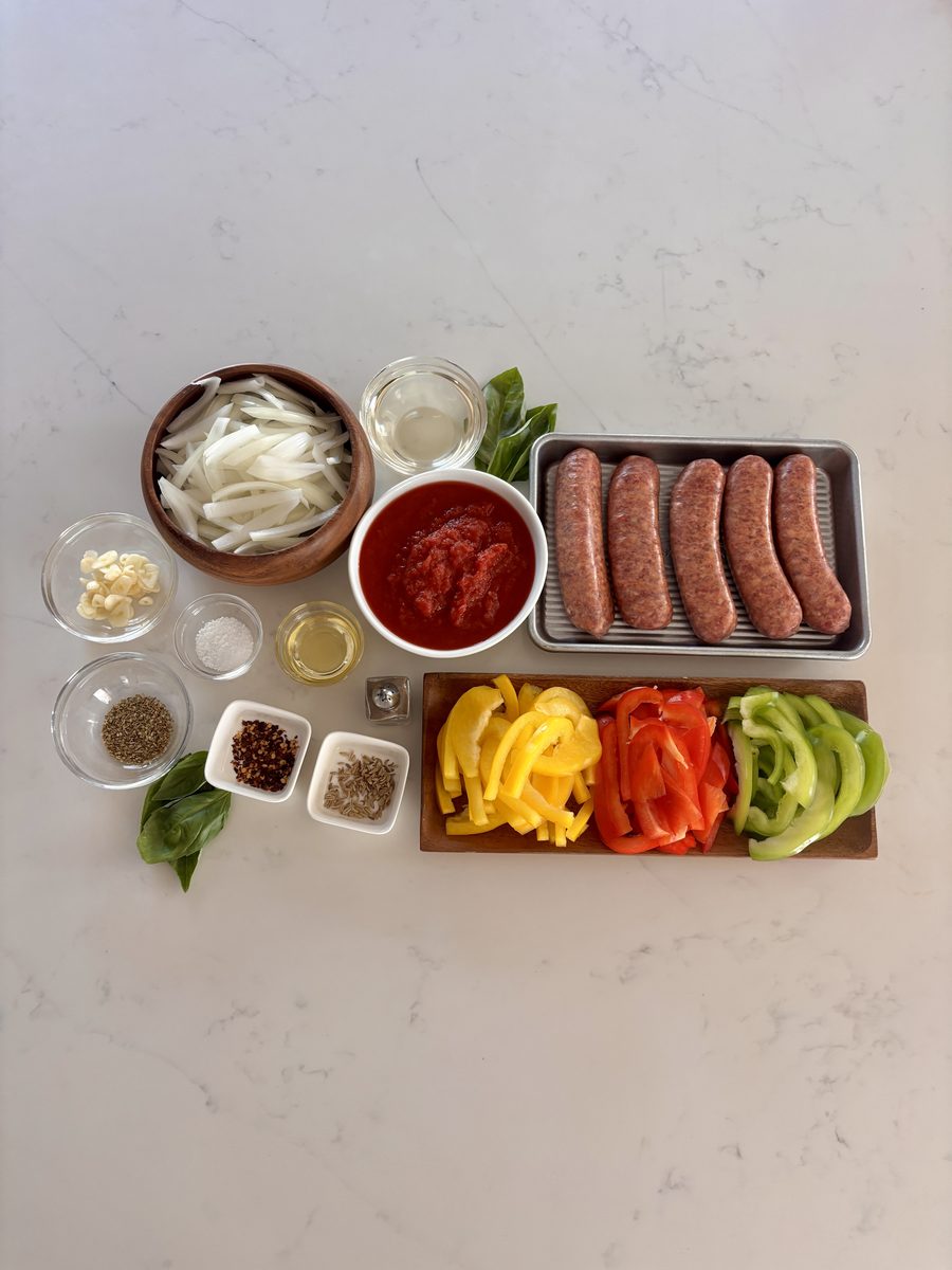 Overhead ingredient layout on white marble: a tray of raw Italian sausage links, a bowl of tomato sauce, sliced yellow, red and gr