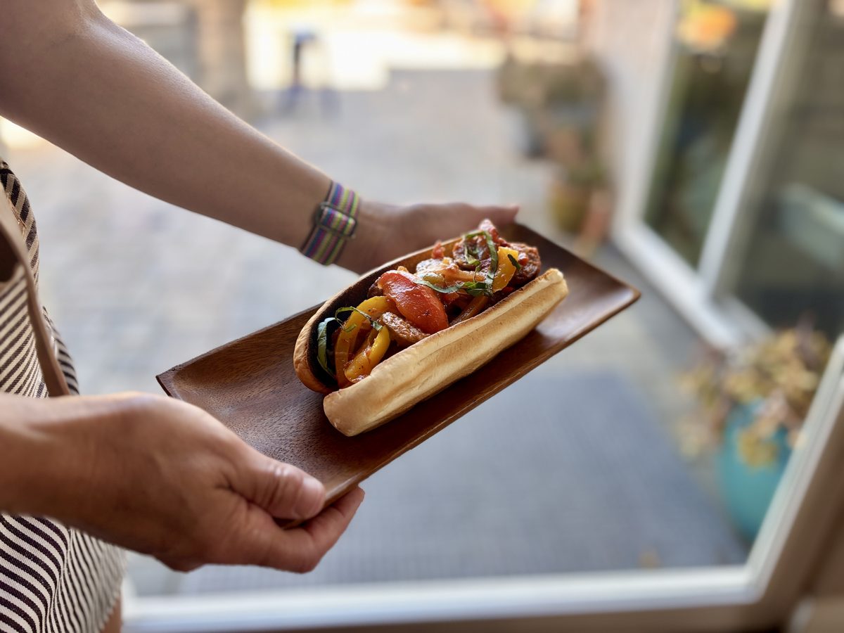 Two hands hold a dark wooden board with a hoagie roll loaded with sausage, red and yellow peppers and onions, outdoor patio backgr