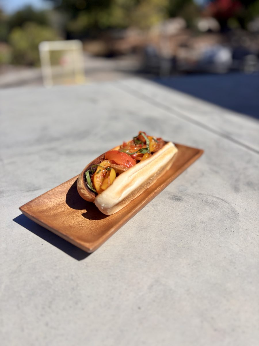 A sausage-and-peppers sandwich on a long roll sitting on a dark wooden serving board on a grey concrete outdoor counter.