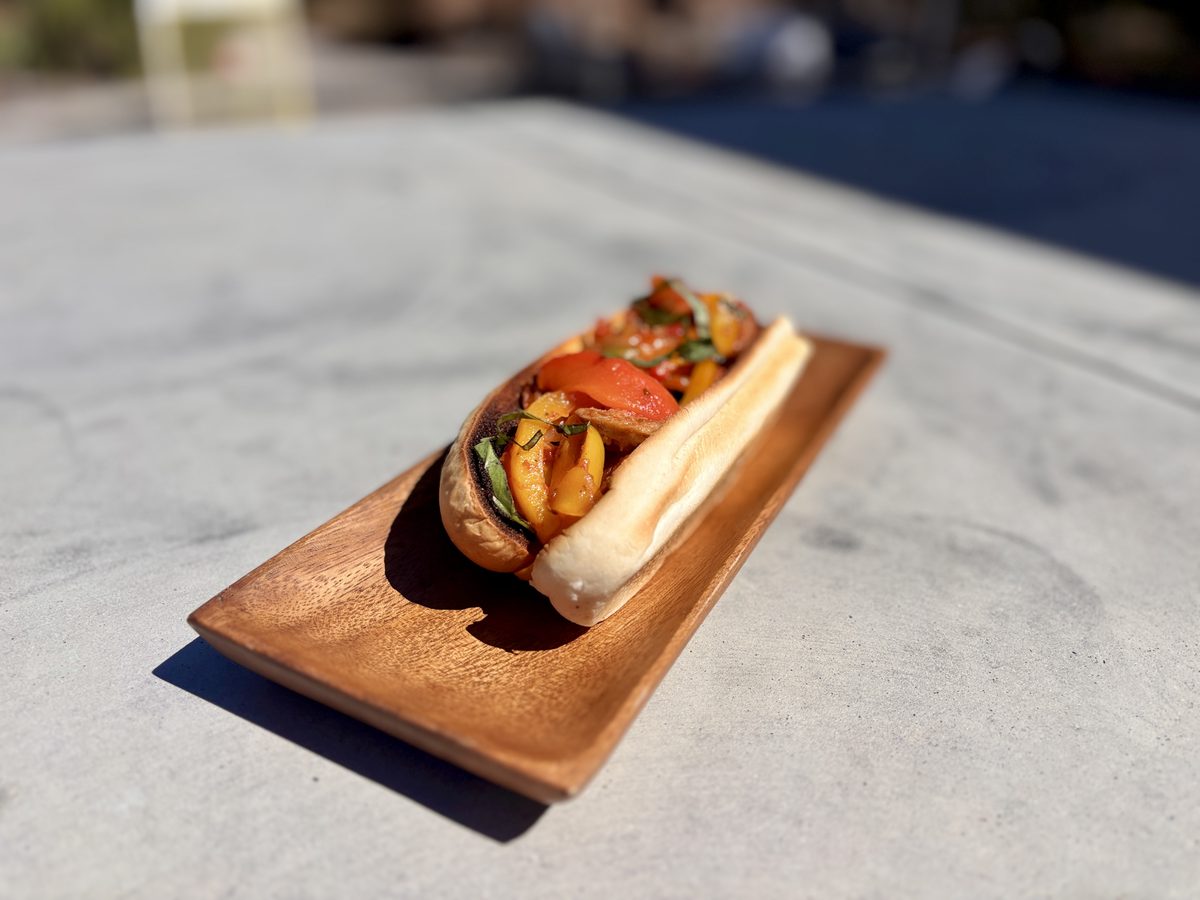A long roll on a narrow wooden serving board filled with browned sausage, peppers and onions, on a marble counter.