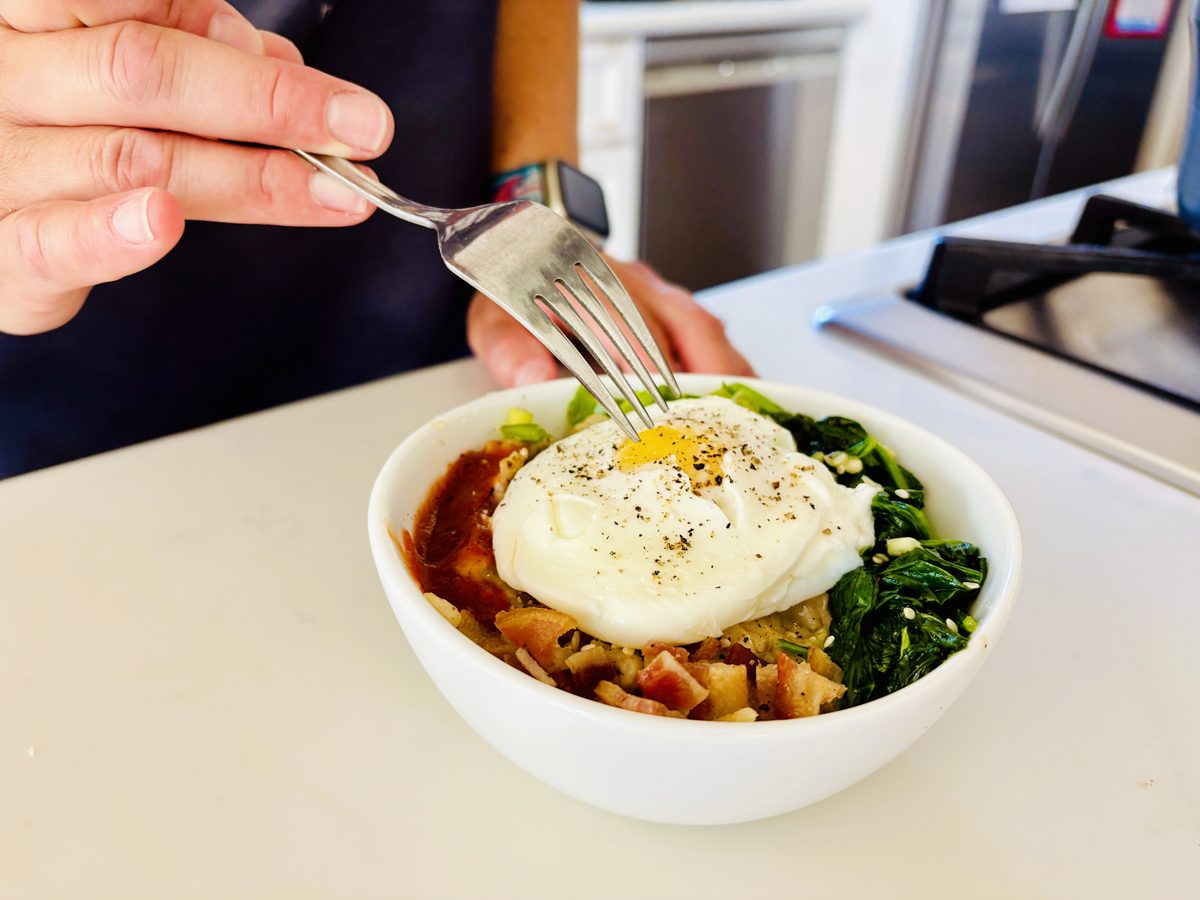 A white bowl of savory grain/oatmeal topped with a poached egg, wilted spinach, diced browned cubes (ham or mushroom, not certain)
