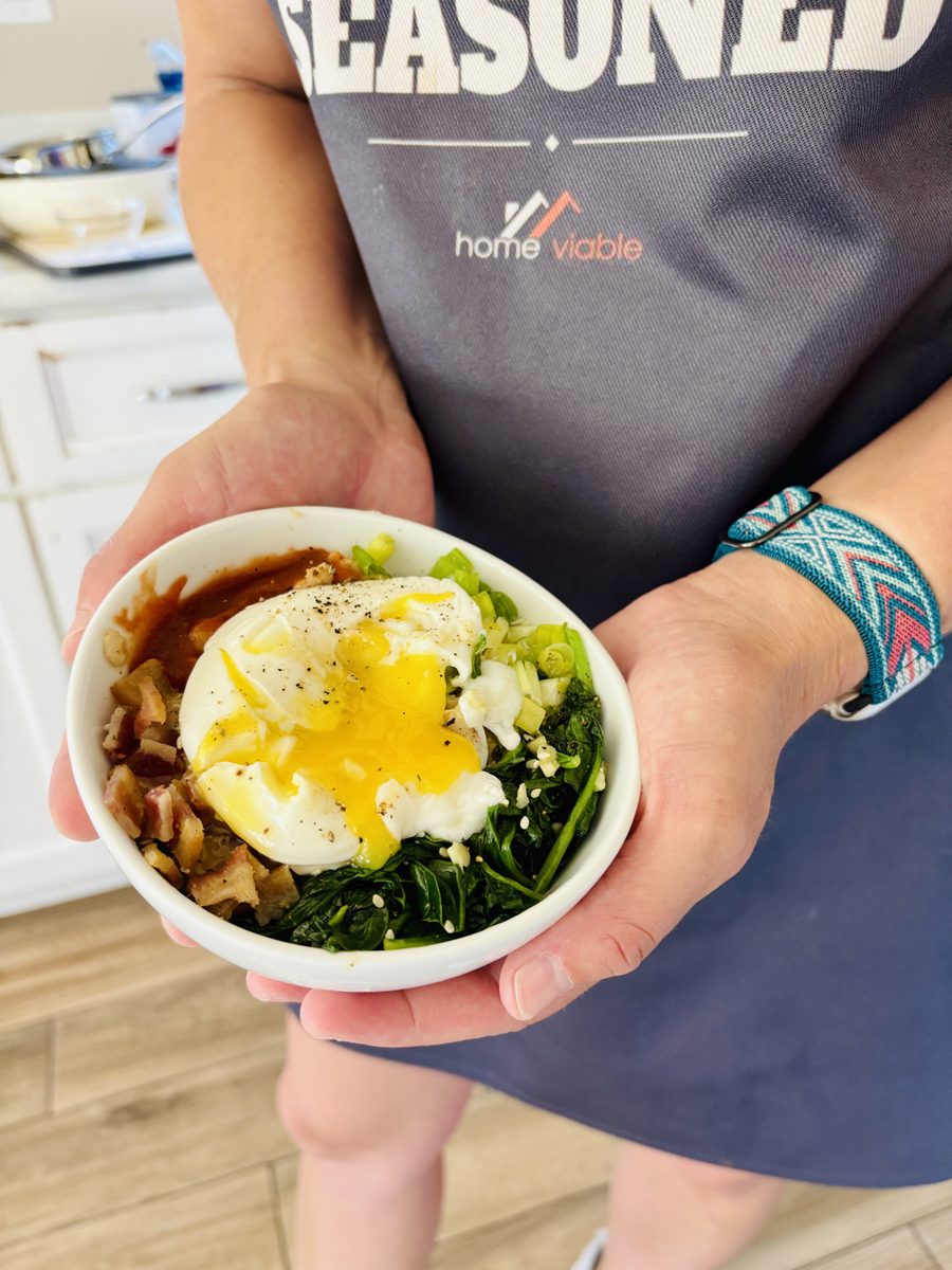 A woman in a dark apron holds a small white bowl of savory oatmeal with a broken poached egg running yolk over spinach, feta crumb