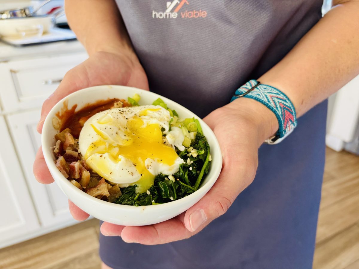 Two hands cradle a white bowl of savory oatmeal with a runny poached egg, spinach, feta and brown diced pieces, held against a gre