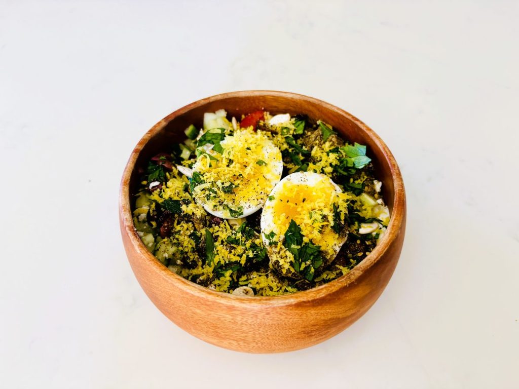 Overhead of a wooden bowl on a white surface holding dark leafy greens topped with two halved boiled eggs and a scattering of bright yellow crumbled topping.