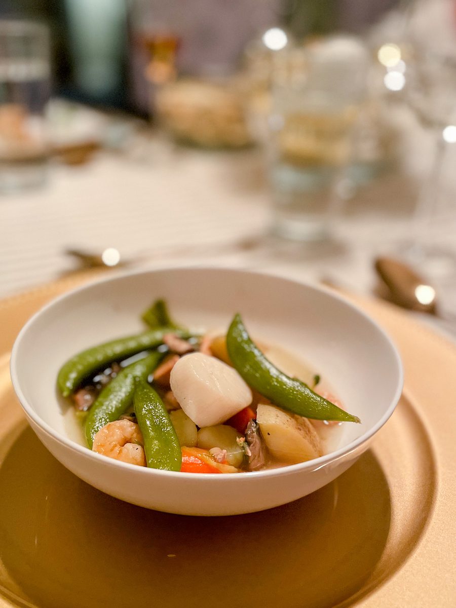 A shallow white bowl of seared scallops and shrimp with sugar snap peas, potatoes and red pepper in broth, on a linen-covered rest