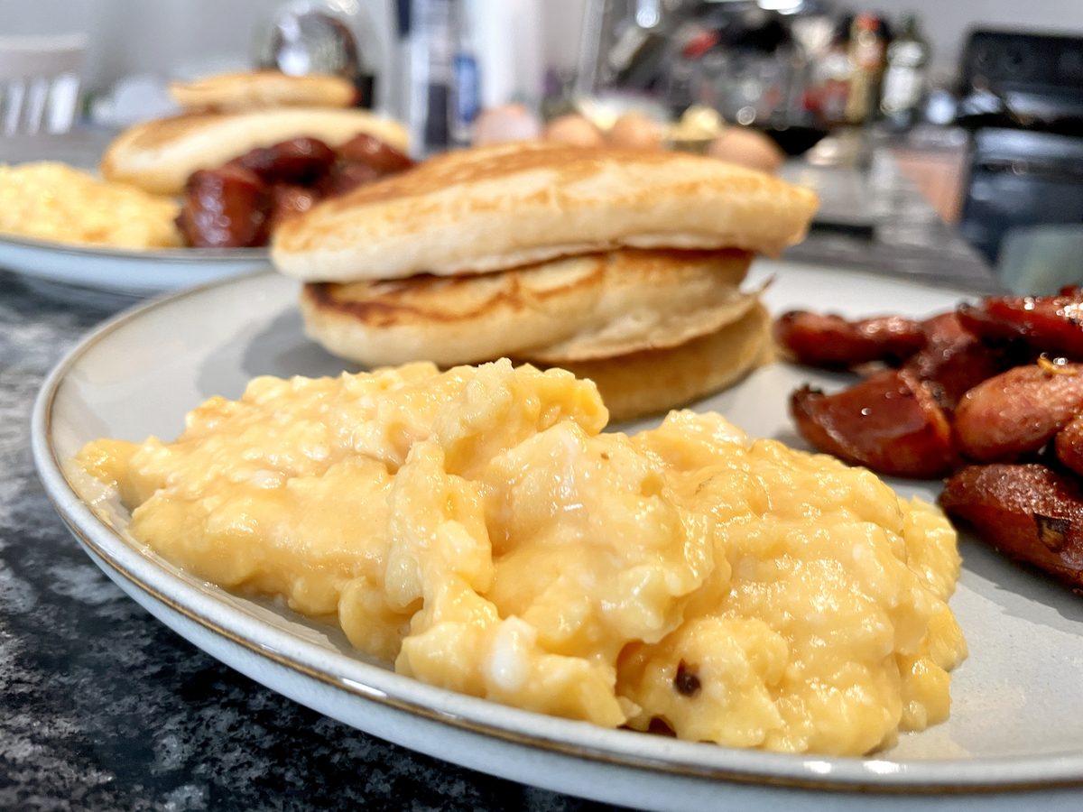 A gray stoneware plate of soft scrambled eggs with pancakes and browned sausage links, shot close on a dark granite counter with a
