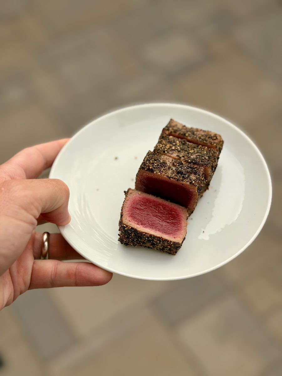 A hand holds a small white plate of sliced pepper-crusted seared ahi tuna showing rare pink centers, outdoors over a stone patio.