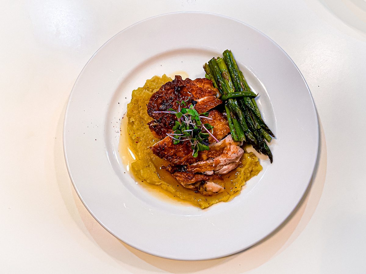 A seared salmon fillet with flaking pink flesh over golden squash puree with roasted asparagus spears and microgreens on a white r