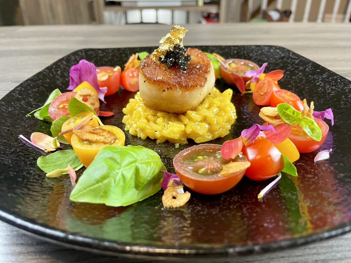 A single seared scallop topped with caviar sits on saffron risotto ringed by halved heirloom cherry tomatoes, basil and edible flo