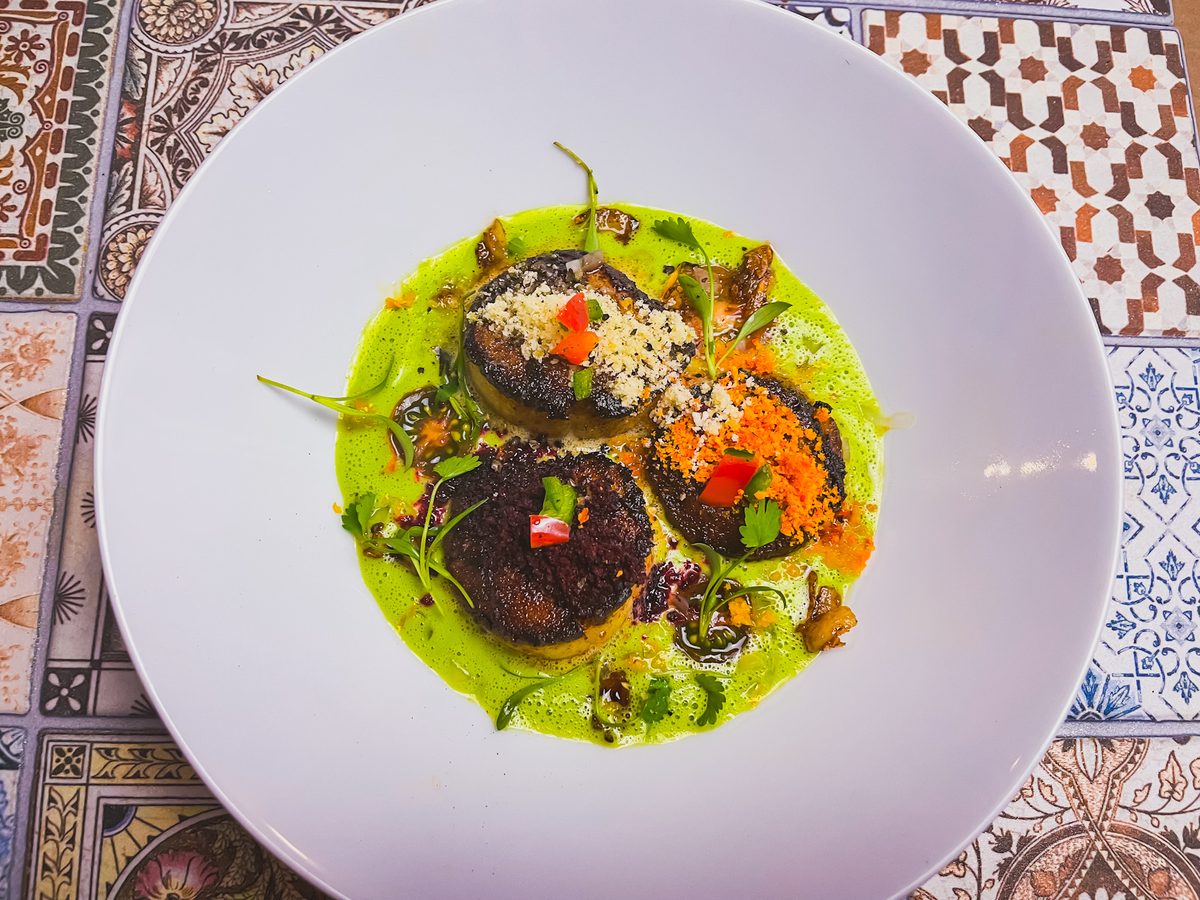 Three heavily charred dark round medallions topped with breadcrumbs, carrot and herbs sit in a pool of bright green sauce in a whi