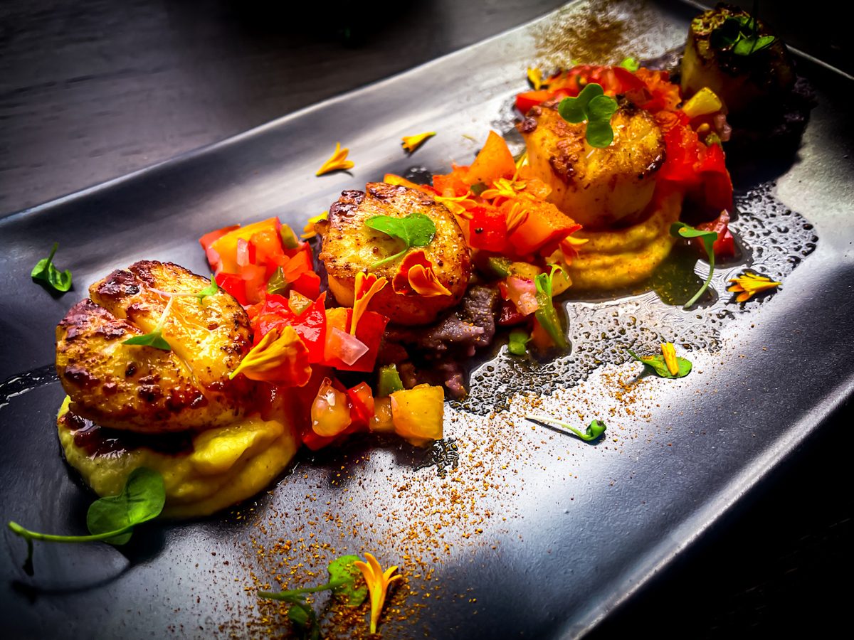 A dark rectangular plate with three seared scallops topped with a bright tomato and pepper relish, dotted with sauce and micro her