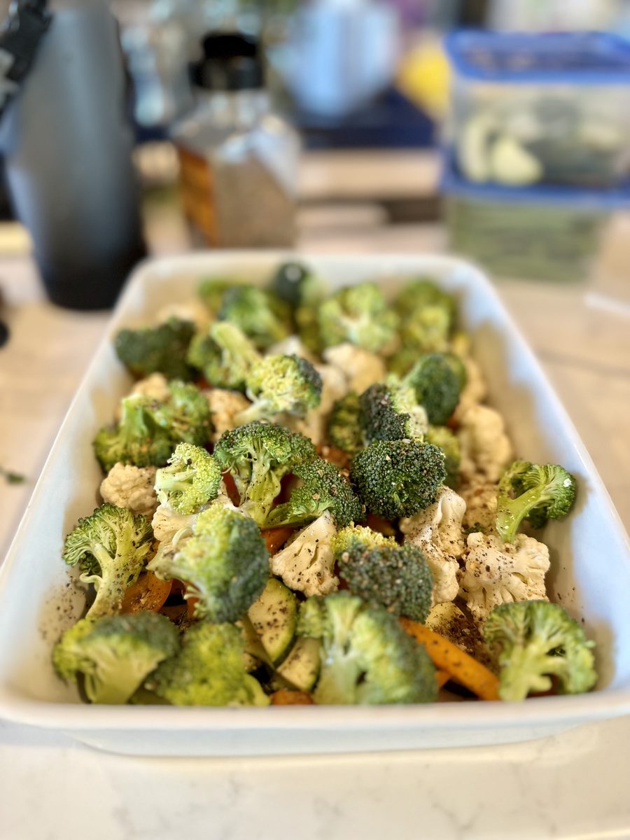 Raw seasoned broccoli, cauliflower and carrot sticks piled in a white rectangular baking dish on a cluttered kitchen counter befor