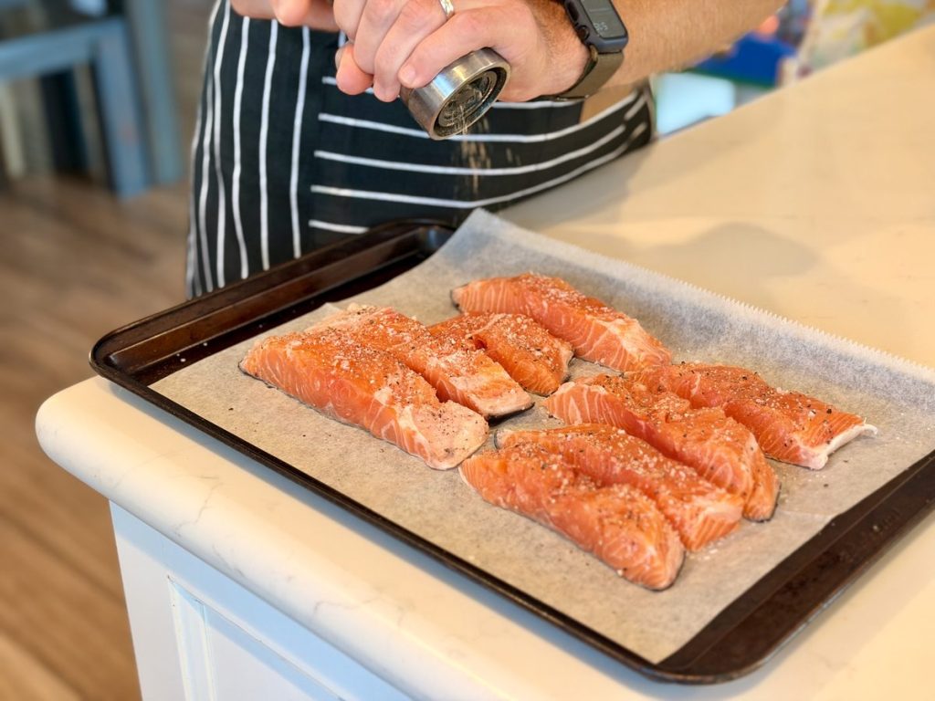 A person in a striped apron grinds seasoning over raw salmon fillets laid on parchment on a sheet pan at a kitchen island; only torso and hands are in frame.
