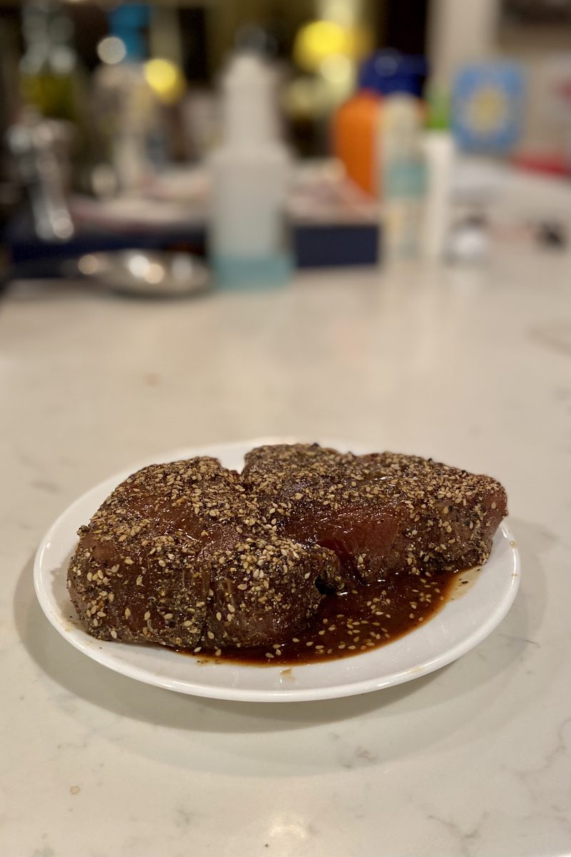 Two sesame-and-pepper-crusted raw tuna pieces on a small white plate with dark soy marinade pooled around them, on a dim counter w