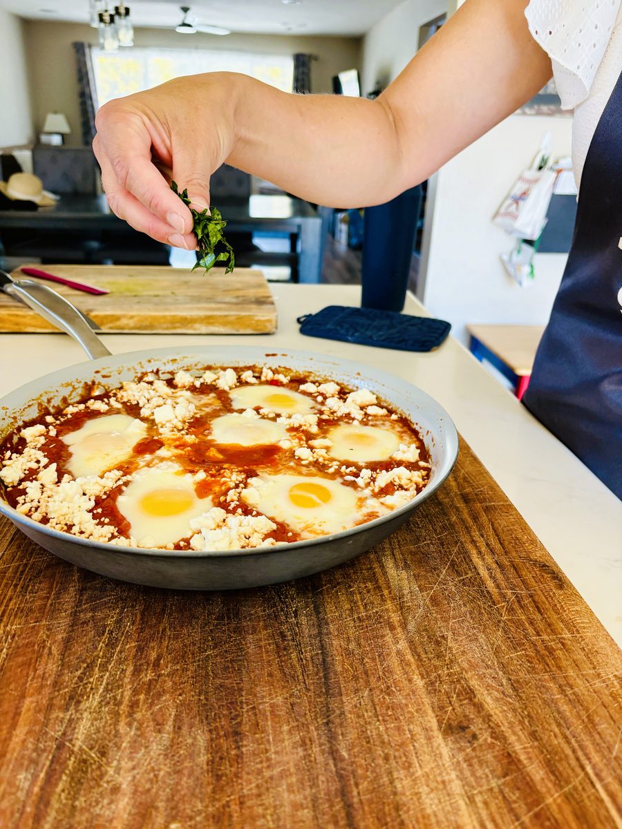 A finished skillet of shakshuka (eggs set in tomato sauce with crumbled feta) resting on a wooden board while a hand sprinkles cho