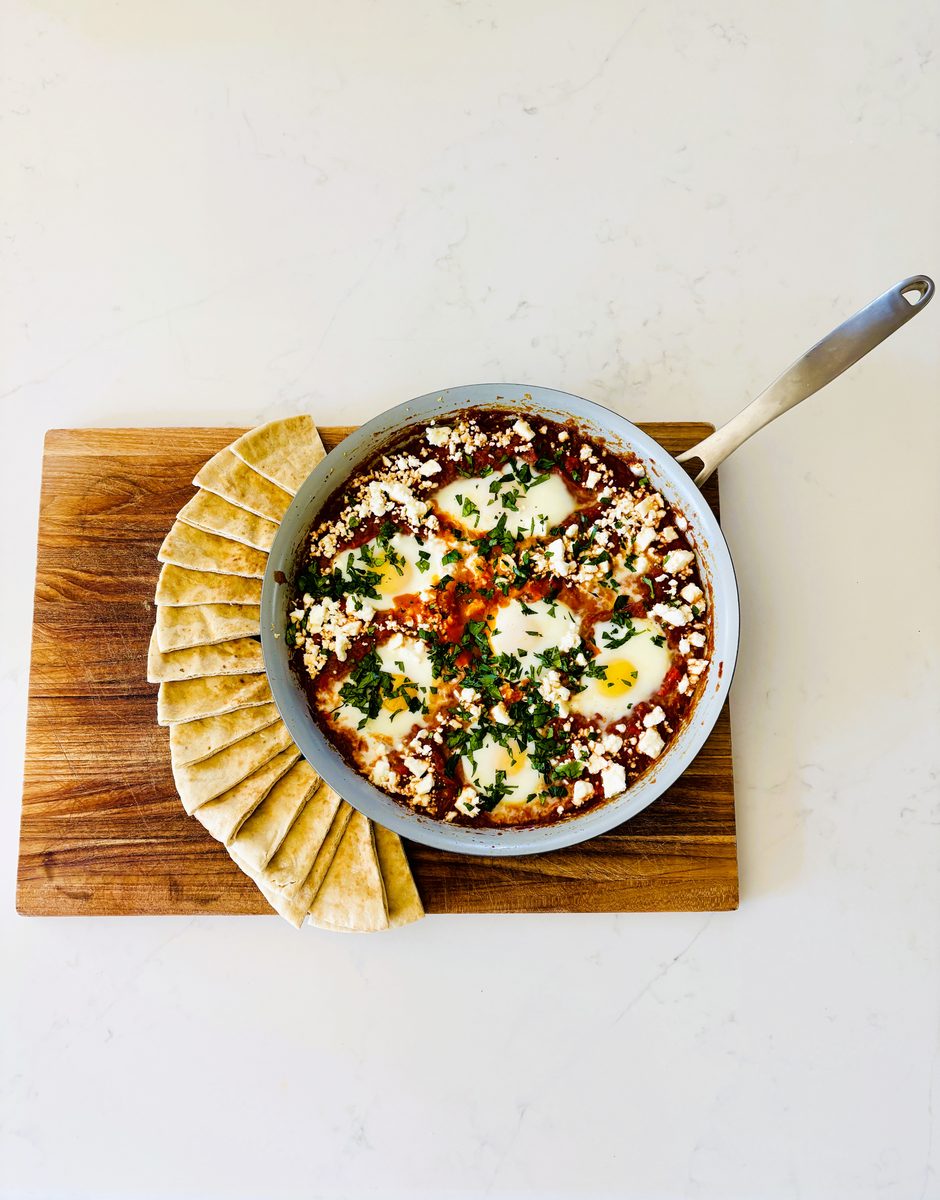 Overhead-angled shot of the finished shakshuka skillet on a wooden board with a fan of pita wedges alongside, on a clean white sur