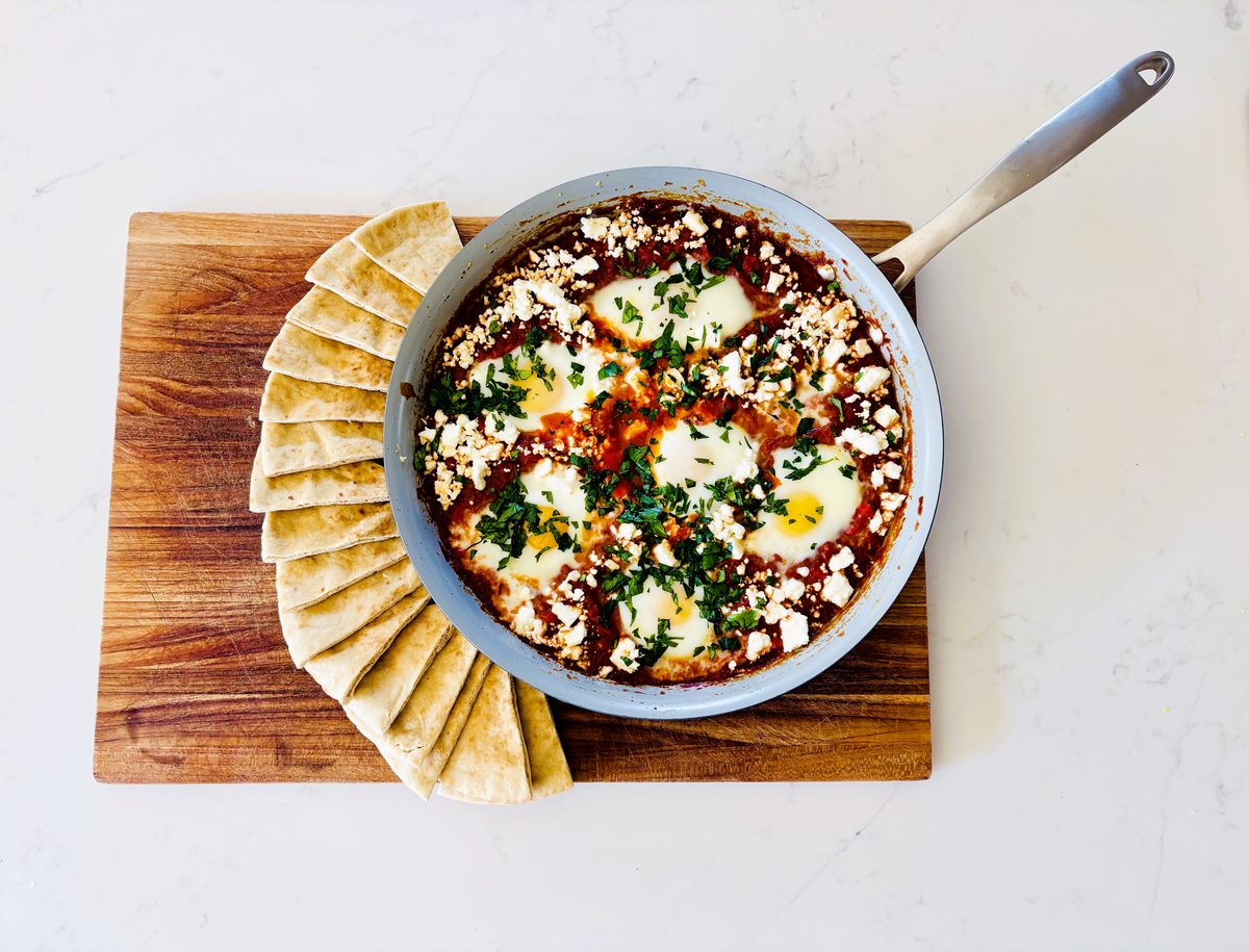 Straight overhead shot of shakshuka in a grey skillet, eggs set in tomato sauce with feta and parsley, on a wooden board ringed by