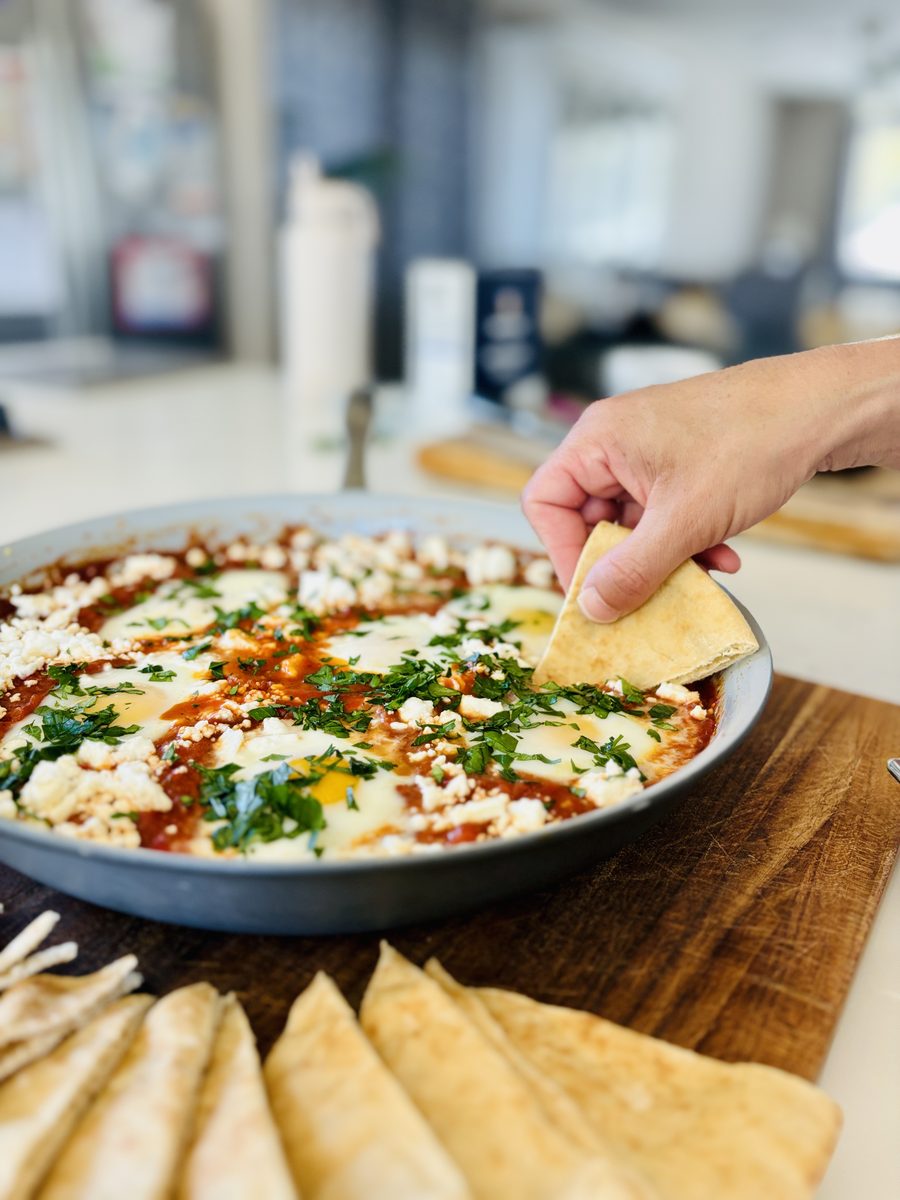 Close low-angle shot of the shakshuka skillet with a hand dipping a pita wedge into the sauce, pita triangles fanned on the board