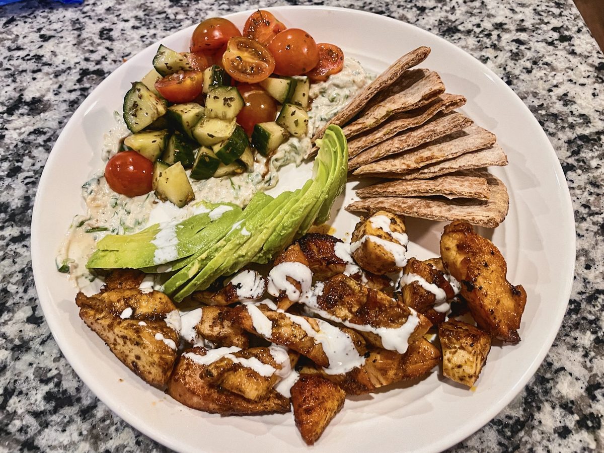 An overhead white plate of shawarma-spiced chicken pieces drizzled with white sauce, alongside rice, a cucumber-tomato salad, cher