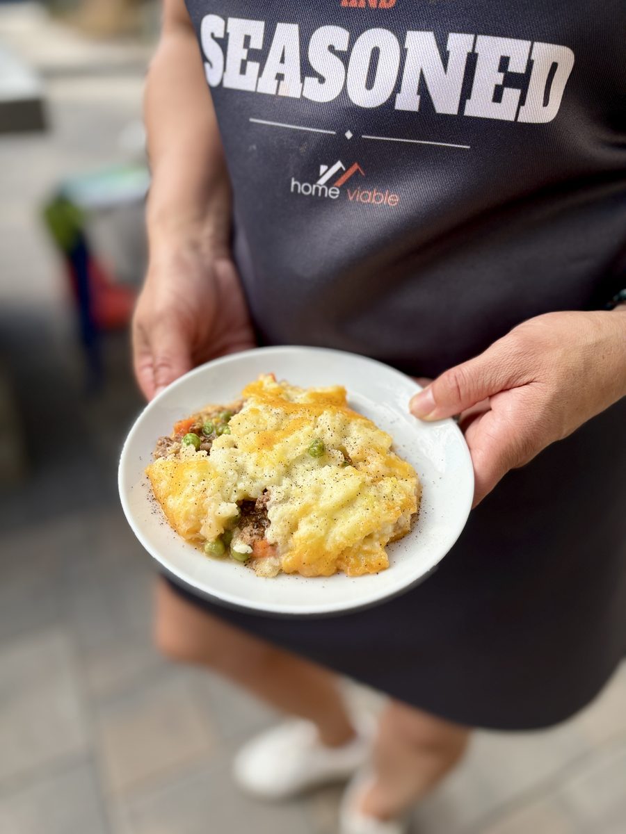 A woman in a dark apron holds a shallow white bowl with a served portion of shepherd's pie showing mashed potato crust over ground