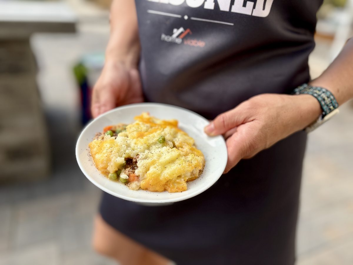 Hands hold a white plate with a scooped serving of shepherd's pie, browned potato topping over ground meat with peas and carrots, 