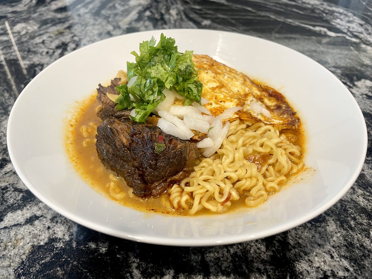 A wide white bowl of ramen noodles in brown broth topped with braised short rib, a halved soft egg and cilantro, shot on dark marb