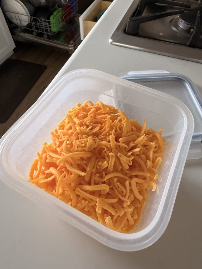 Shredded cheddar cheese portioned into a clear plastic storage container with the lid beside it