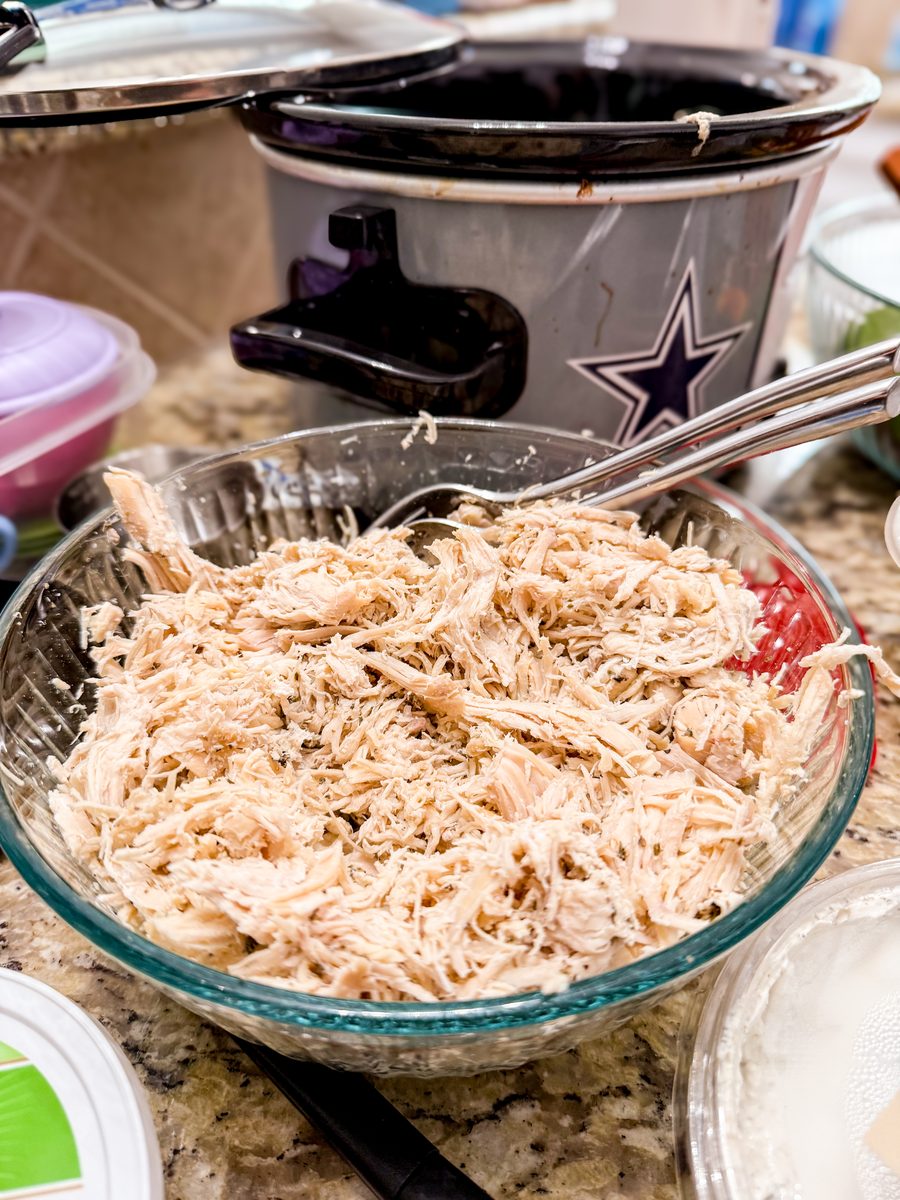A glass mixing bowl heaped with shredded cooked chicken and a serving spoon, sitting on a cluttered counter in front of a slow coo