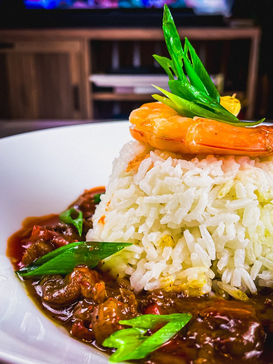 A white plate with a molded mound of white rice crowned by a whole shrimp, surrounded by dark reddish-brown stew with snow peas an