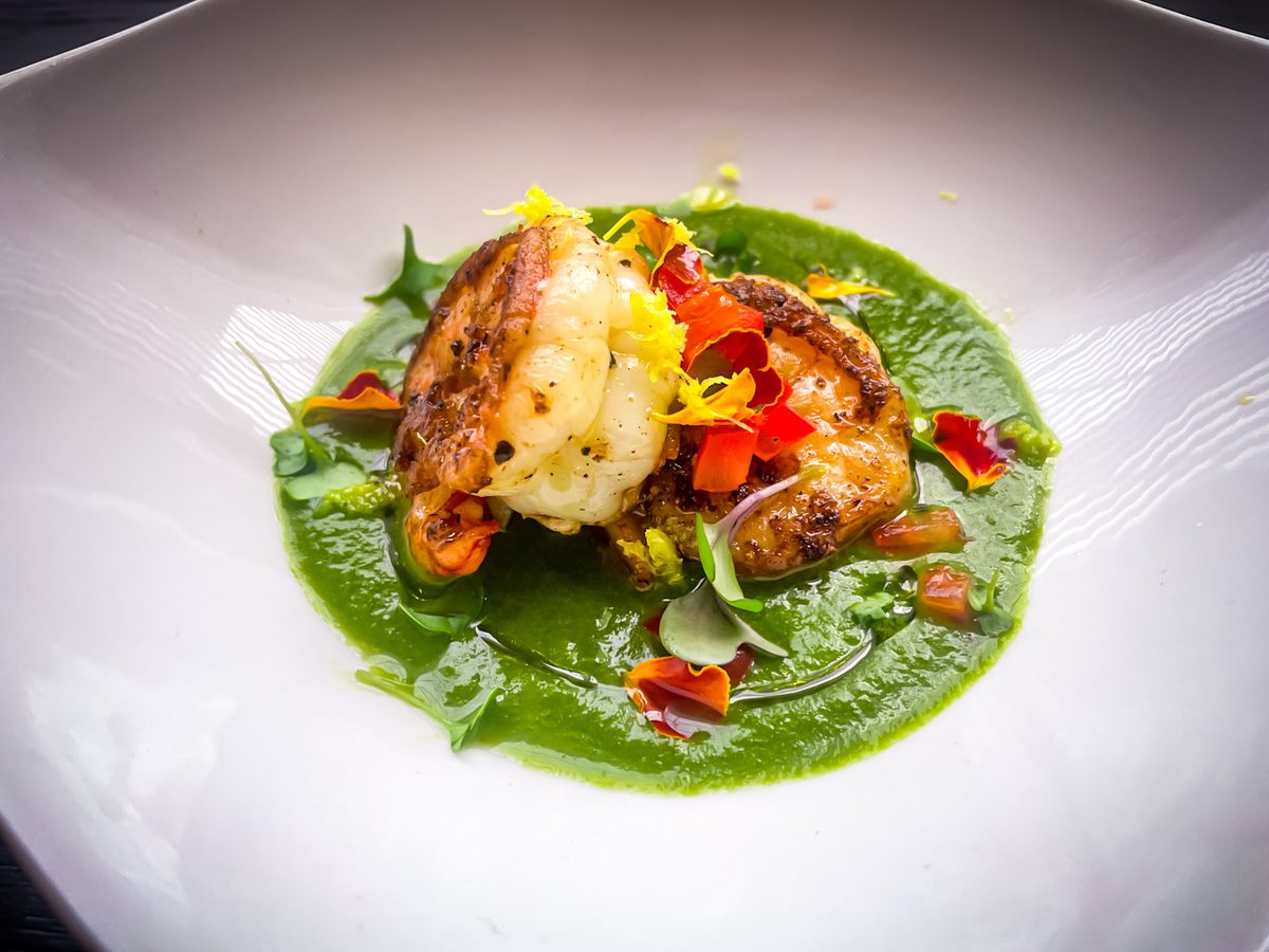 A white plate with grilled shrimp stacked on a swipe of bright green pea puree, garnished with herbs and edible flowers.