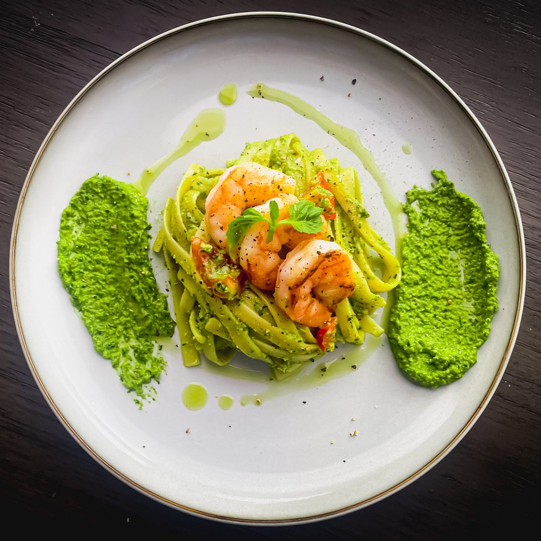 A twirl of pesto-coated pasta topped with shrimp, flanked by two quenelles of bright green pea puree on a white plate.