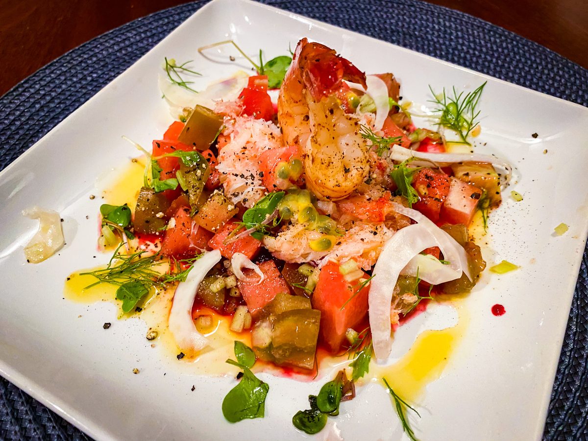 Grilled shrimp over a chopped watermelon, tomato and shaved onion salad dressed with olive oil and dill, on a white square plate a