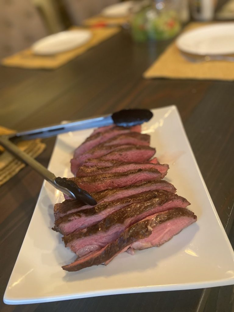 Sliced grilled flank steak fanned out on a white platter with serving tongs