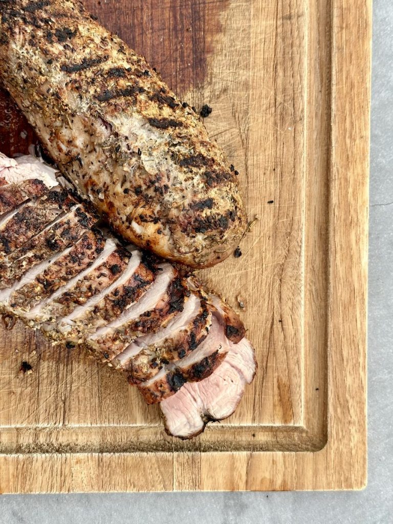 Two grilled pork tenderloins on a wooden board, one whole and one sliced into thick medallions showing juicy pink centers.