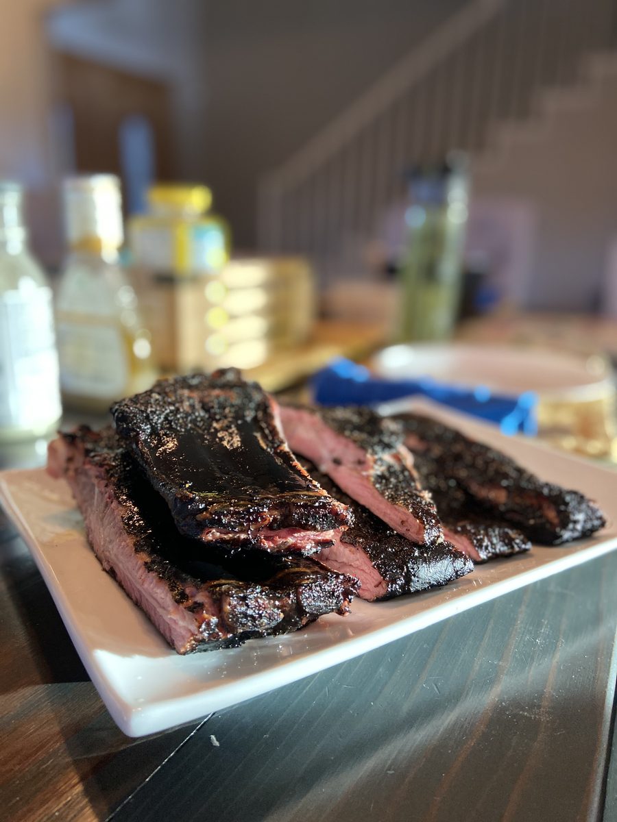 Sliced smoked brisket with a dark bark and pink smoke ring fanned on a white rectangular platter, with blurred bottles and a clutt
