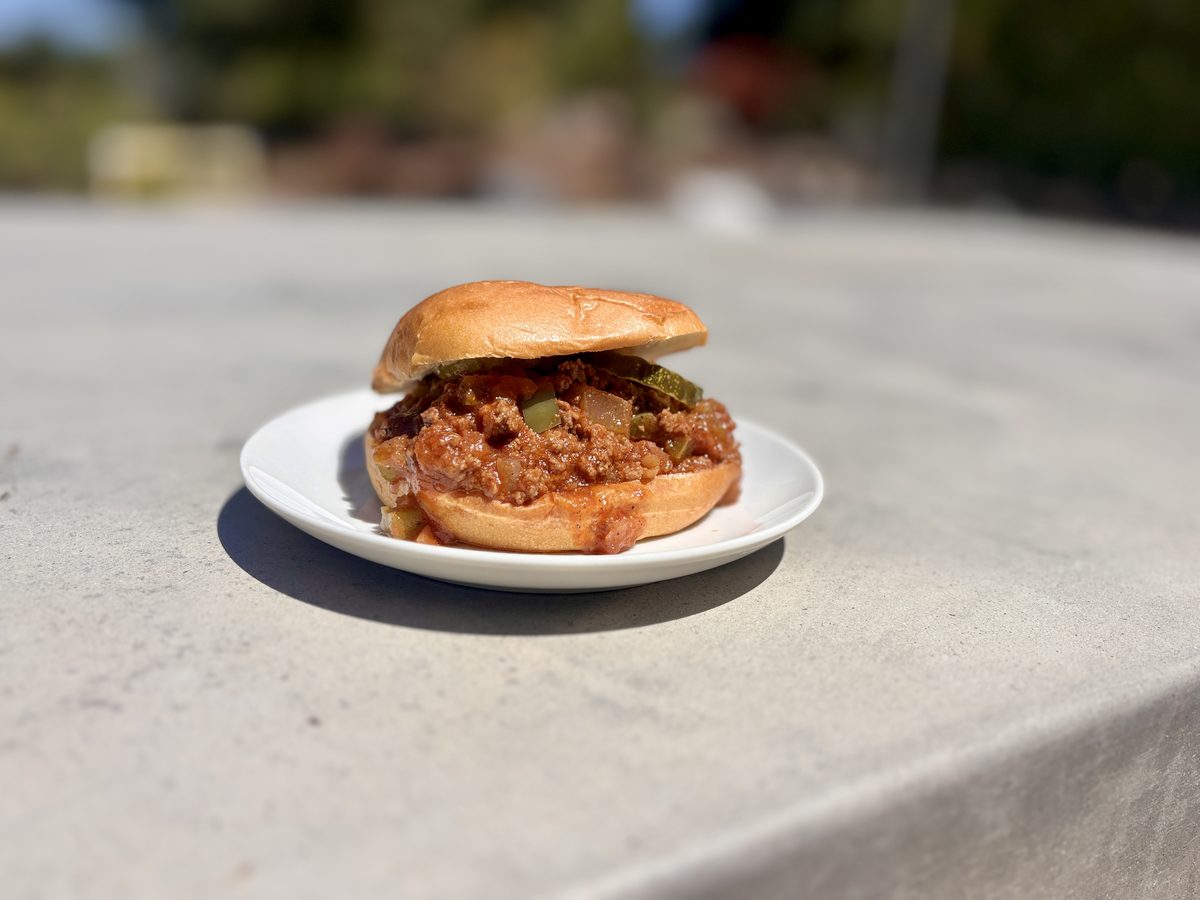 A small white plate on a marble counter with a single sandwich on a soft golden bun overflowing with saucy brown ground meat.