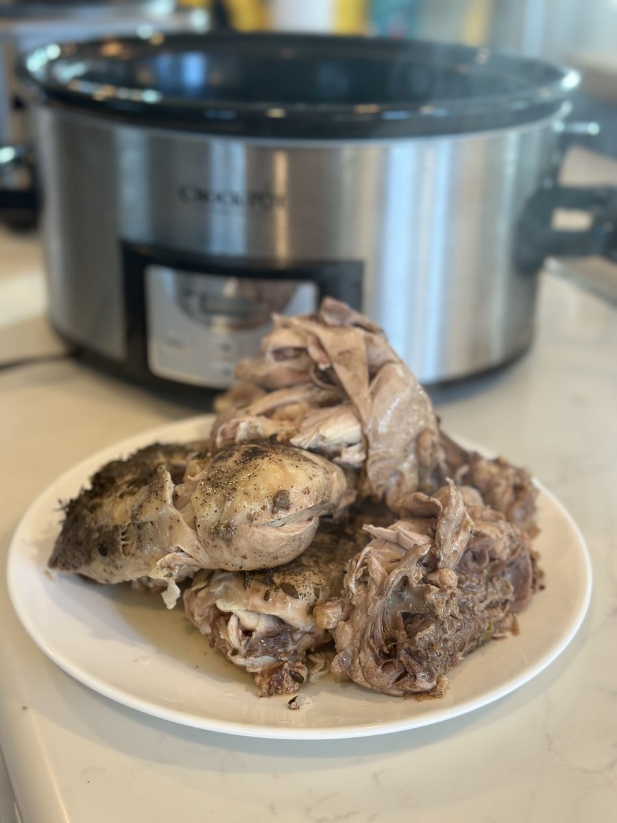 A pile of cooked, pulled bone-in chicken pieces on a white plate on a counter directly in front of a stainless slow cooker.