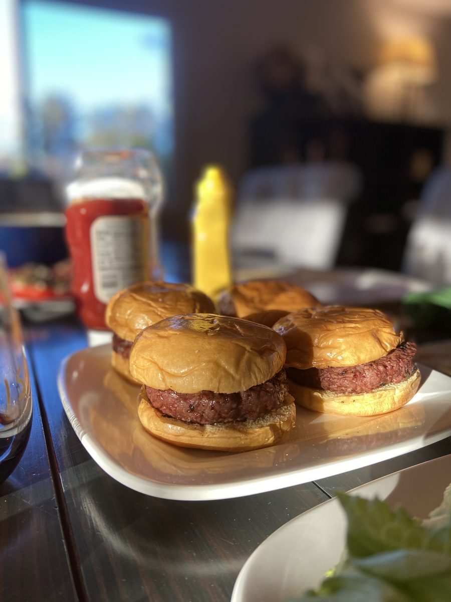 Four smash burger sliders on brioche buns stacked on a white plate in a dim room, with blurred ketchup and mustard squeeze bottles