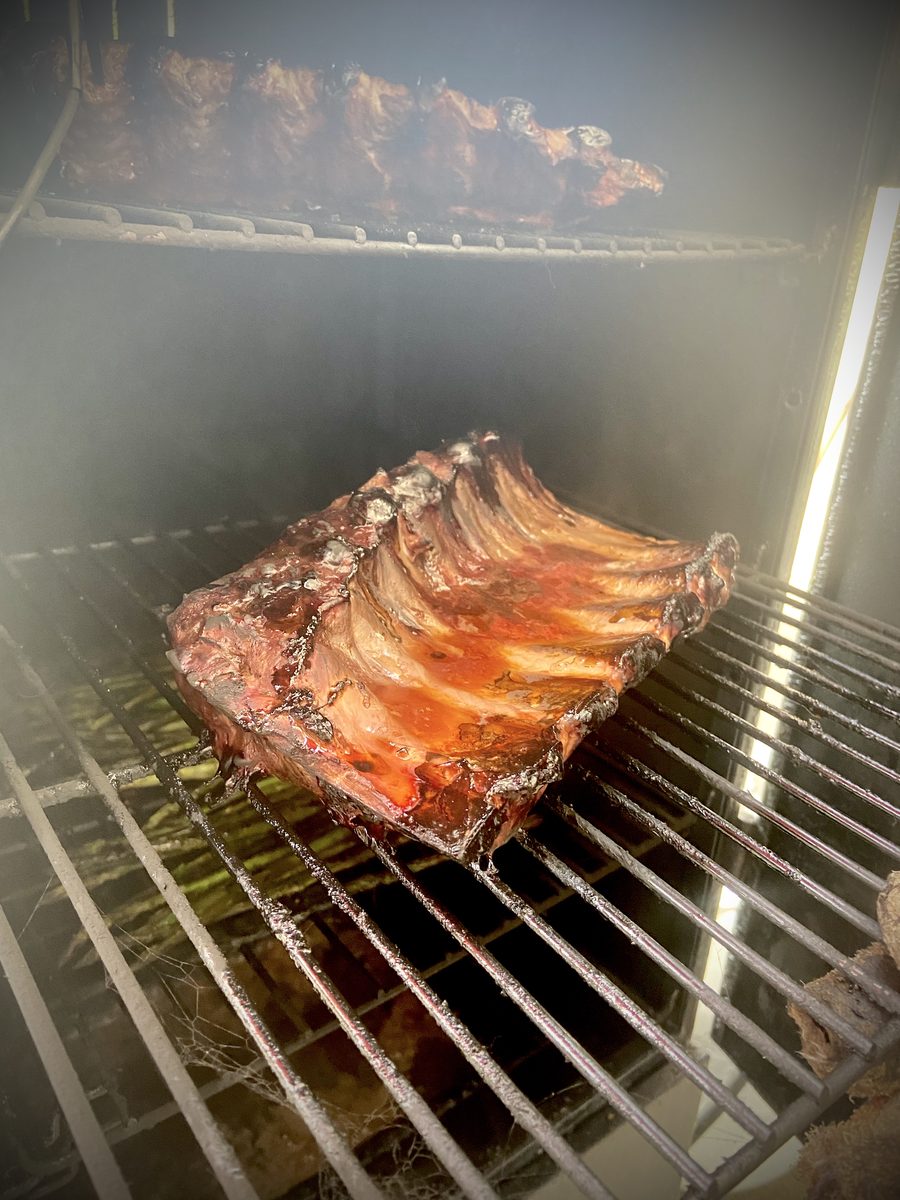A rack of glazed barbecue ribs sitting on the grate inside a smoker, smoke haze visible around the racks.
