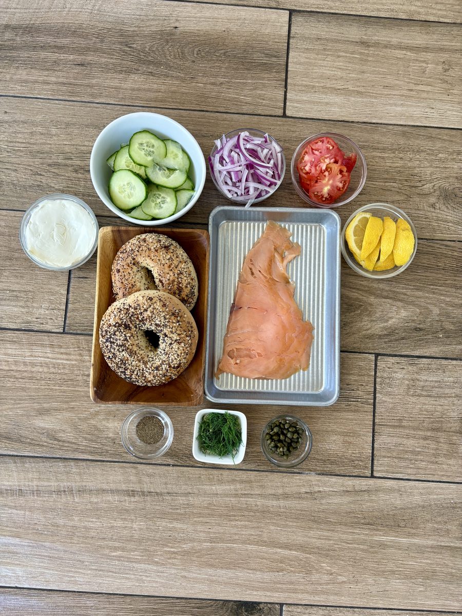 Overhead flat lay on a wood floor of bagels, a slab of smoked salmon in a metal tray, cream cheese, sliced cucumber, red onion, to