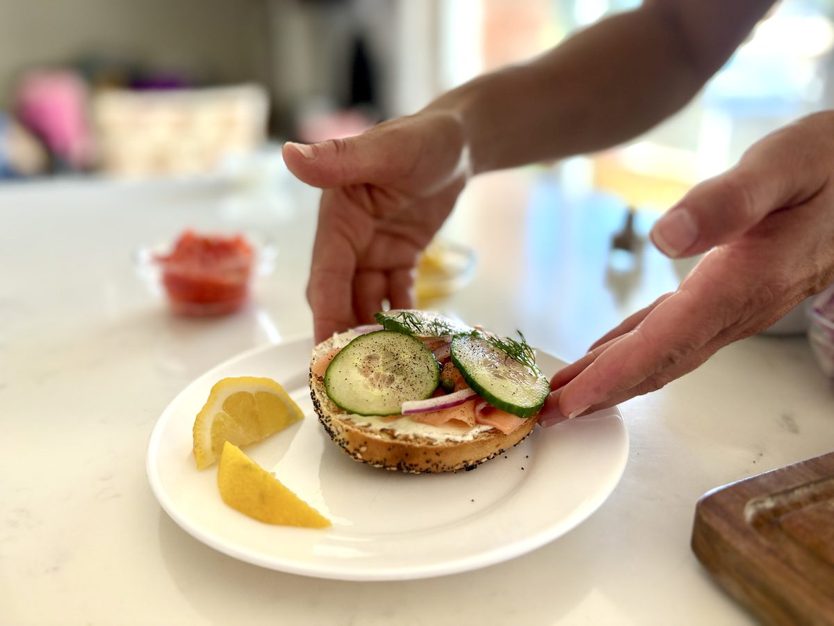 Hands laying cucumber slices onto an open-faced everything bagel with cream cheese, smoked salmon, red onion and dill on a white p
