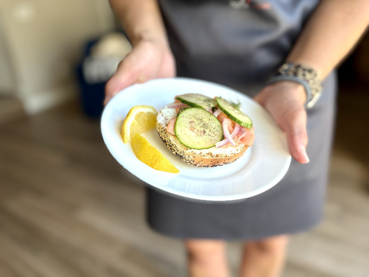 A woman holds out a white plate with an open-faced smoked salmon bagel topped with cream cheese, cucumber, red onion and dill, plu