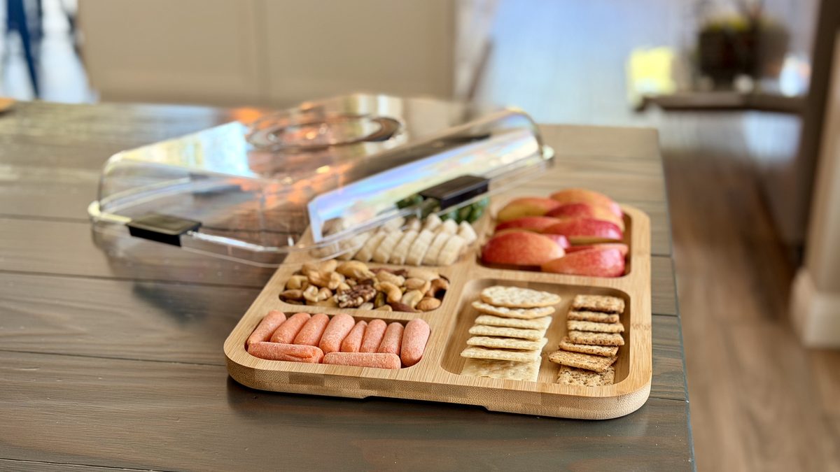 A compartmented bamboo snack tray on a table holding two kinds of crackers, walnuts, white cheese slices, apple slices and pink sl