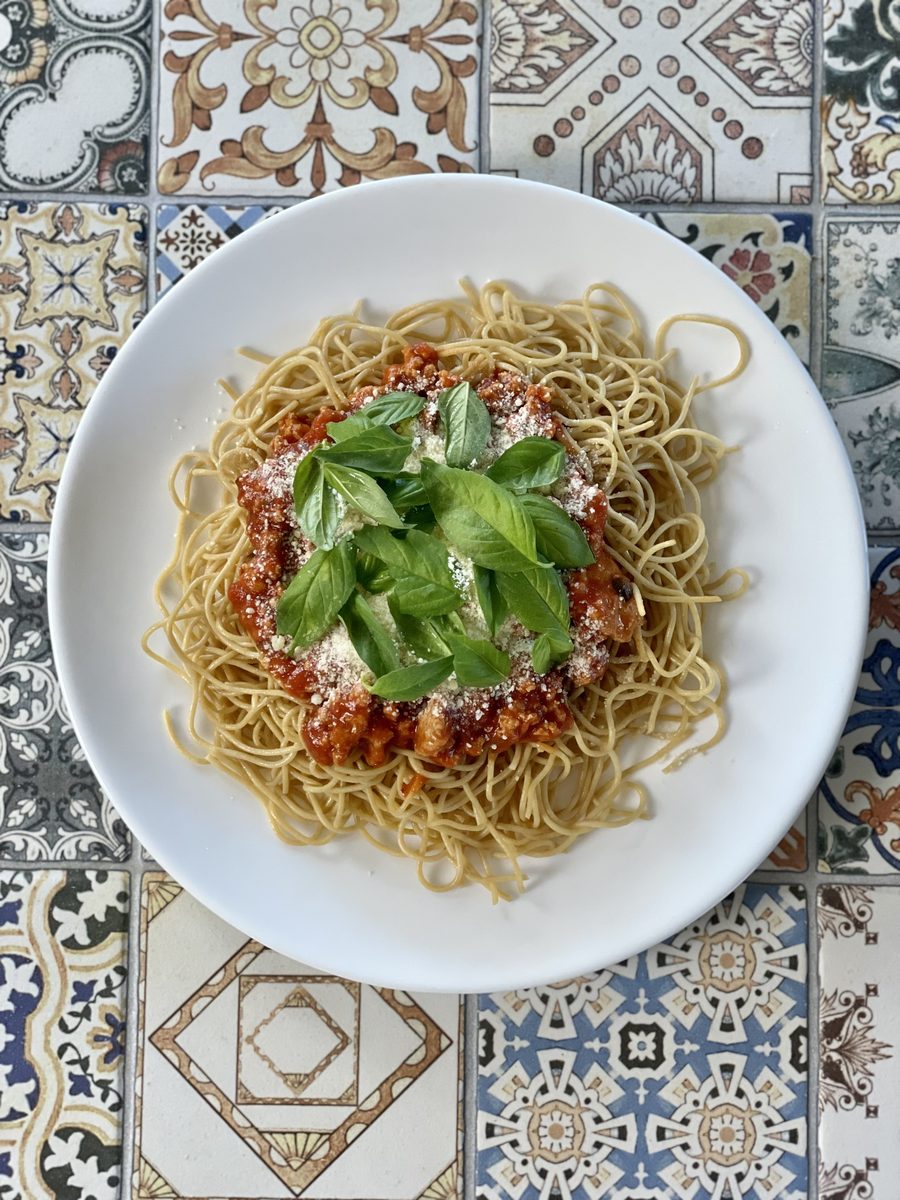 Overhead white bowl of spaghetti with marinara, grated parmesan and a crown of fresh basil leaves, on ornate blue and tan patterne