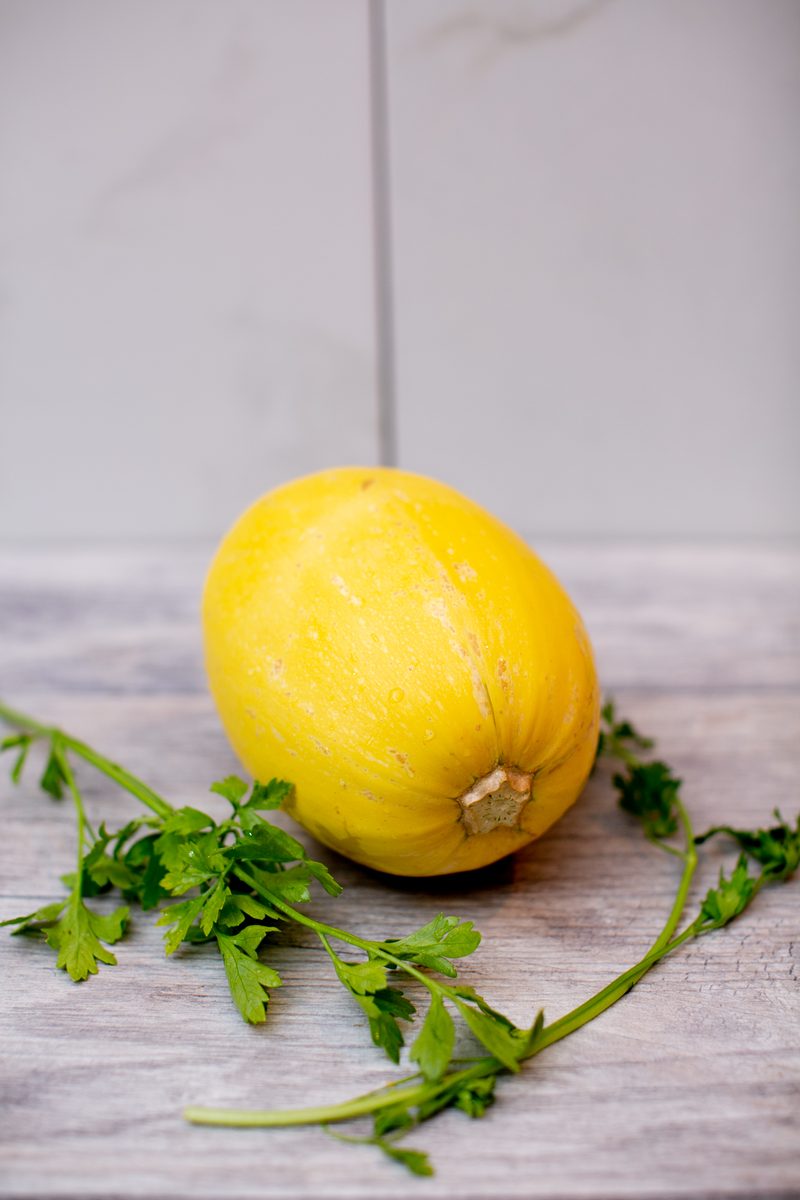 A whole uncut yellow spaghetti squash resting on a pale wood surface with a sprig of fresh parsley beside it.