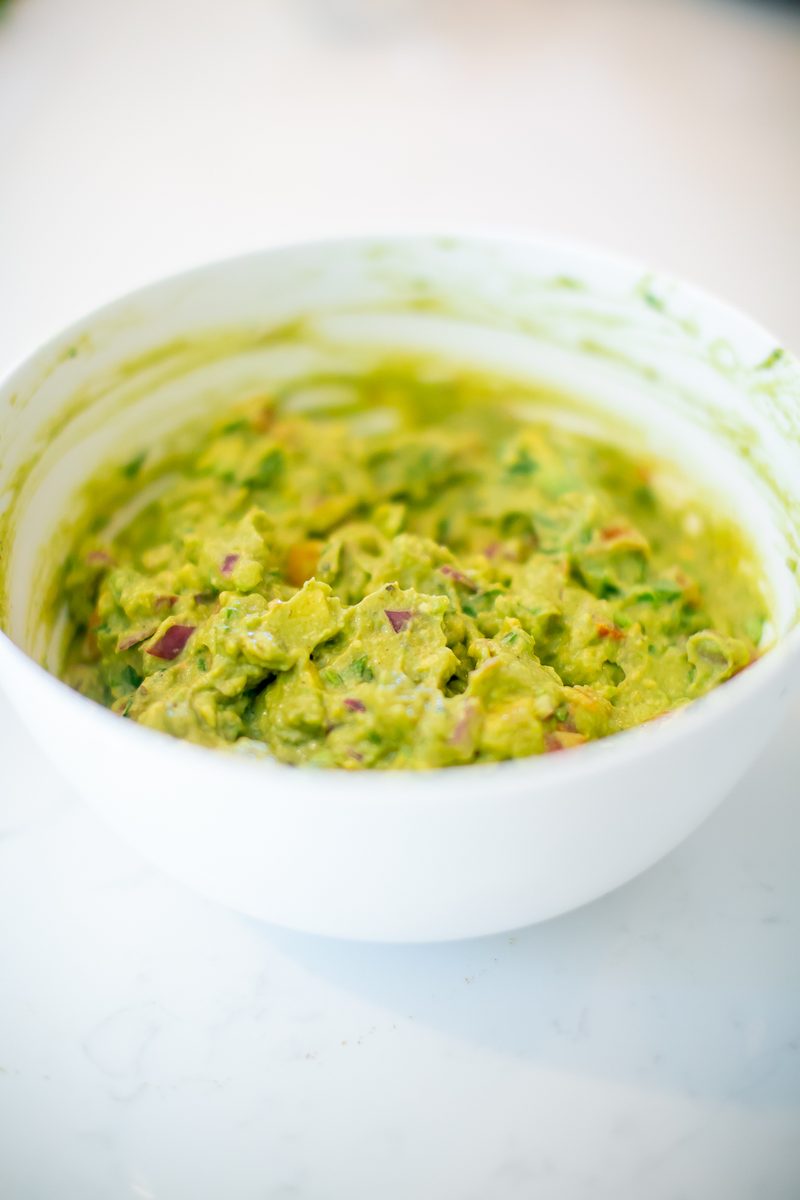 A white bowl filled with chunky green guacamole flecked with red pepper and herbs.