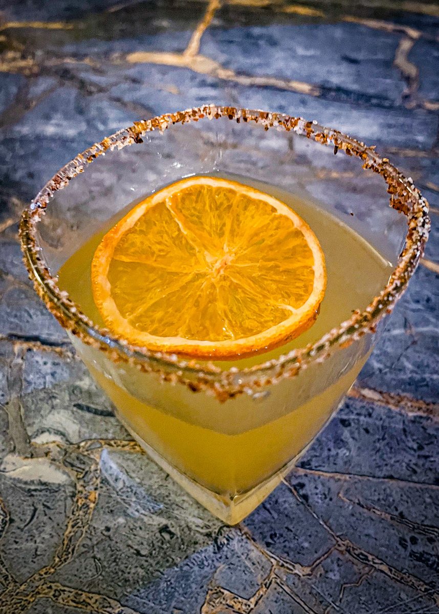 A chili-salt-rimmed coupe of orange margarita garnished with a dried orange wheel, standing on a dark blue slate slab.